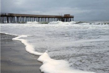 Surf crashing on the beach in New York, with a brooding grey sky serving as a backdrop to a pier leading out into the ocean into the background.