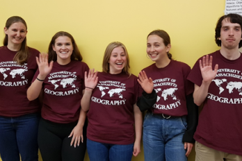 Five UMass Geography students standing in front of a yellow wall, wearing red t-shirts with U-Mass Geography logo, smiling waving at camera.