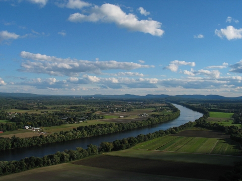 View looking south from Mt. Sugarloaf of Connecticut river winding its way towards bottom left of image, bordered on both sides by green farm fields. The distinctive peaks of the Holyoke range are in the background.